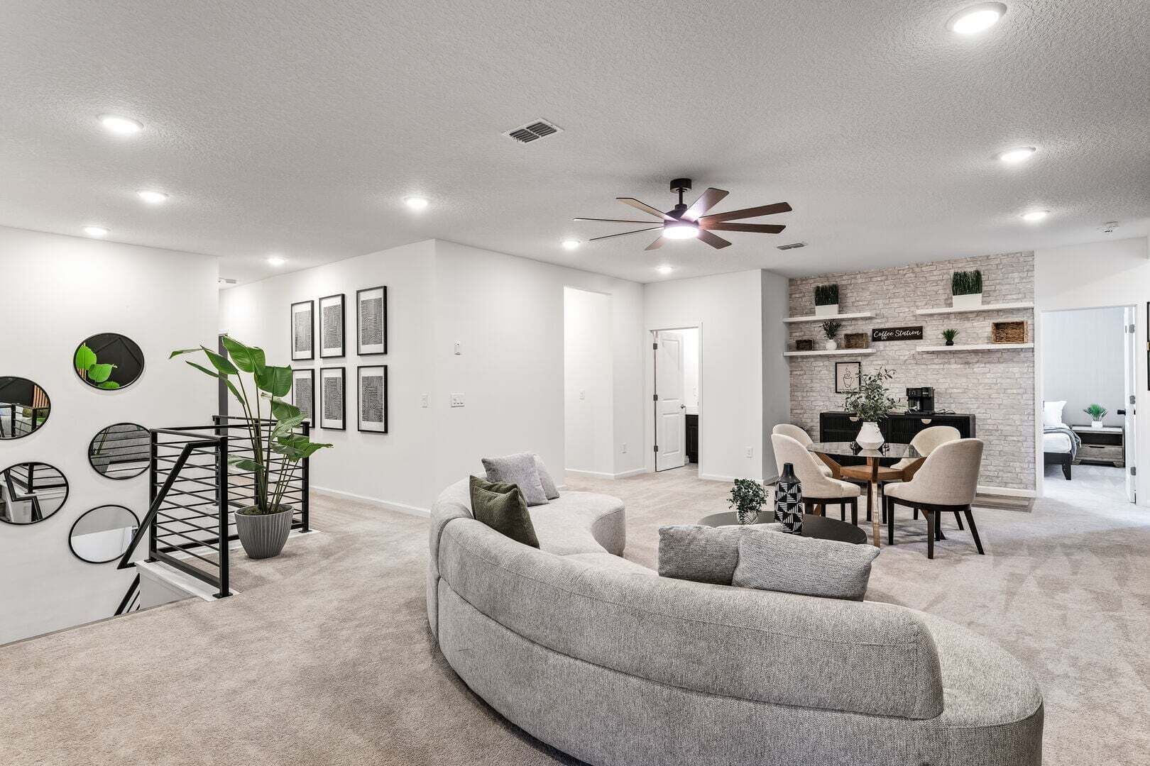 A modern living room with a grey sofa, a dining area with a table and chairs, a staircase, and a kitchen area with a brick wall and shelves.