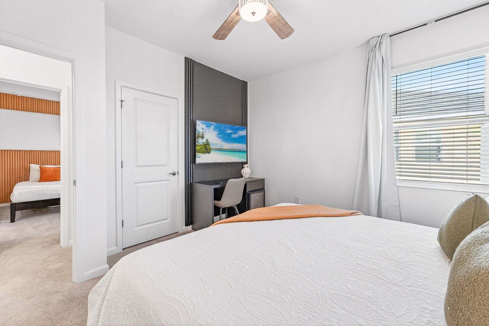 A bedroom with a bed, a desk, a chair, a television, and a window with blinds.