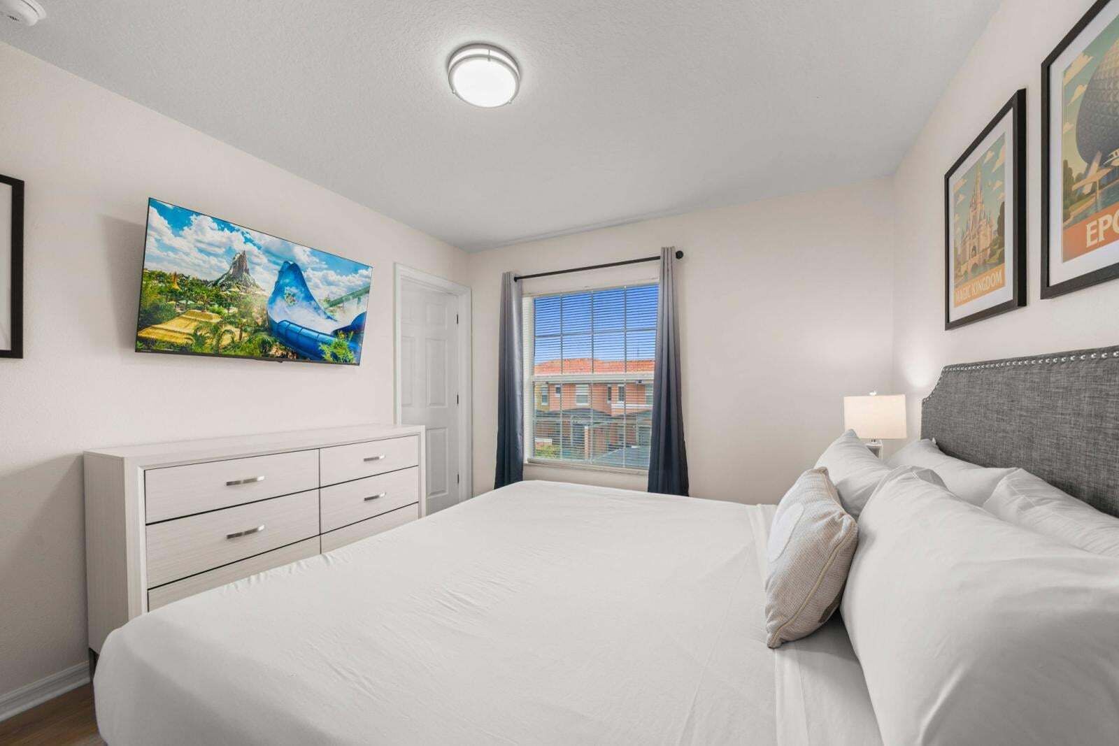 Ein Schlafzimmer mit Bett, Schminktisch, einem an der Wand montierten Fernseher und einem Fenster mit Blick auf Gebäude.