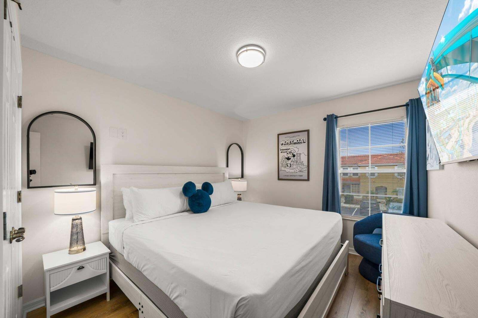 A bedroom with a bed, bedside tables, a mirror, a lamp, a window with curtains, and a wall-mounted picture.