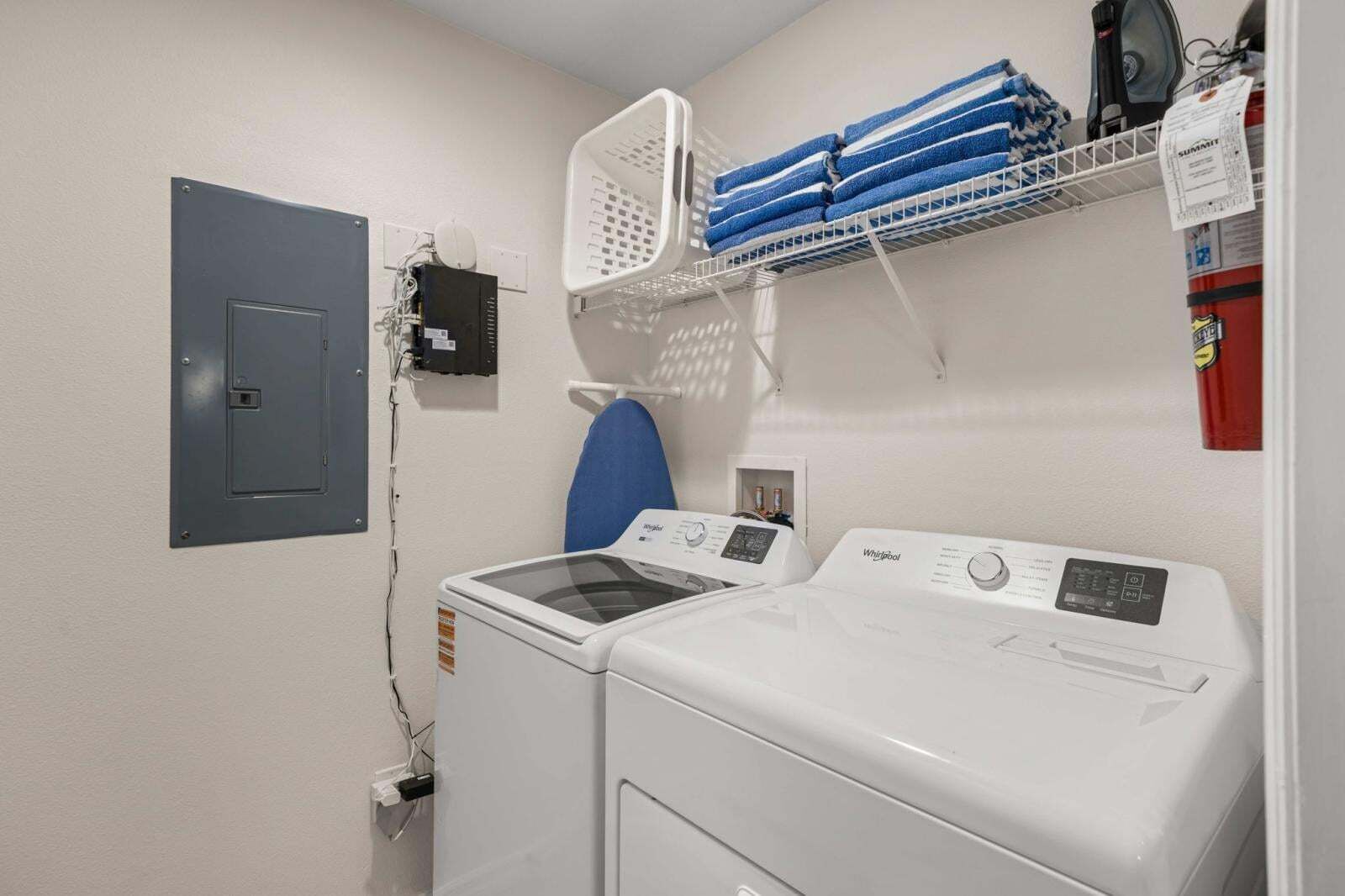 A laundry room with a washing machine, dryer, iron, and a shelf with folded towels.