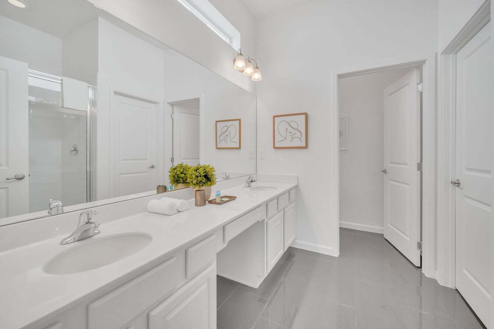 A modern bathroom with a large mirror, two sinks, a shower, and a hallway leading to a door.