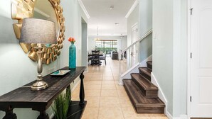 Un pasillo con un espejo decorativo, una mesa con una lámpara, un jarrón con flores y una escalera que conduce a otra habitación.