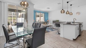 Een moderne keuken met een centraal eiland, een eethoek met een glazen tafel en een zithoek met een bank en televisie.