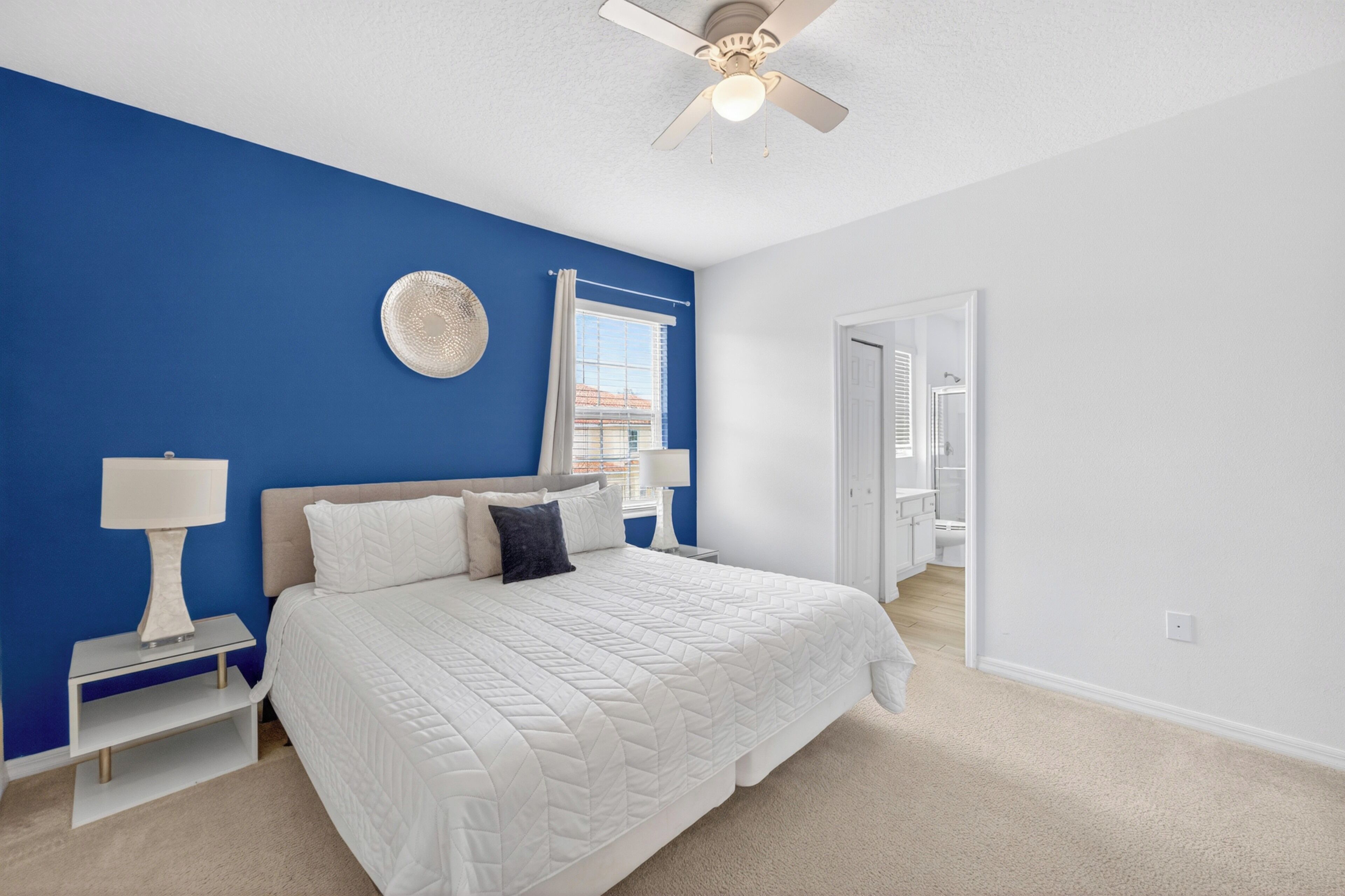 A bedroom with a bed, nightstands, a ceiling fan, a window with blinds, and a door leading to another room.