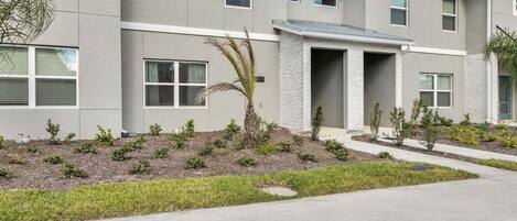A modern multi-story building with a grey facade, a central entrance with a stone archway, and a landscaped front yard with small shrubs and a palm tree.