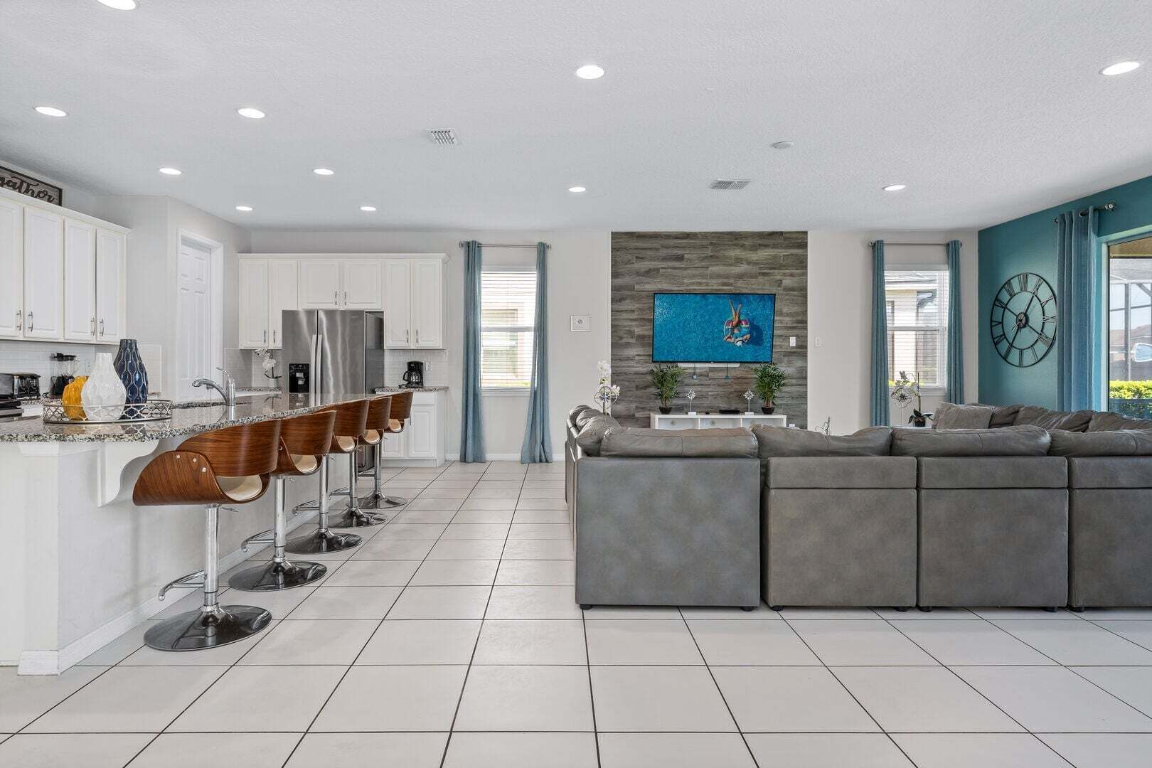A modern kitchen with white cabinets, a bar area, and a large grey sectional sofa in a living area with a stone accent wall and a clock.