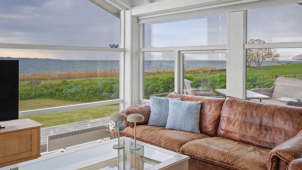 Ein Zimmer mit Ledersofa, einem Glastisch und einem Fernseher auf einer Holzkommode. Große Fenster bieten Blick auf das Meer und die grüne Umgebung.
