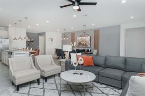 Ein modernes Wohnzimmer mit grauem Sofa, zwei Sesseln, einem Couchtisch, Pendelleuchten und einem Deckenventilator.