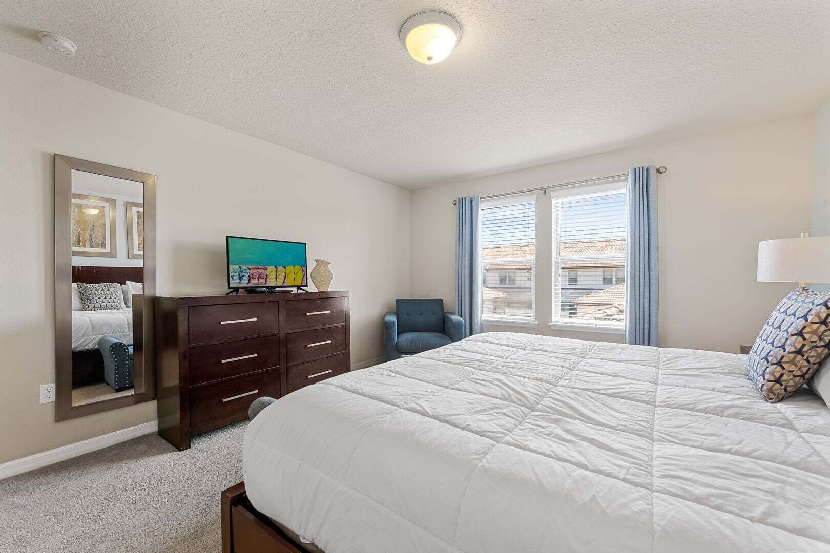 Un dormitorio con una cama grande, un aparador, una silla, un televisor y un ventanal con cortinas.
