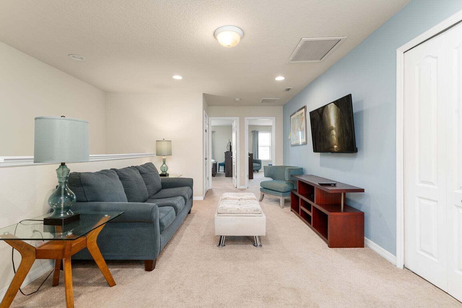 Una sala de estar con sofá, reposapiés, mesa de centro de vidrio, televisor y mueble para televisión.
