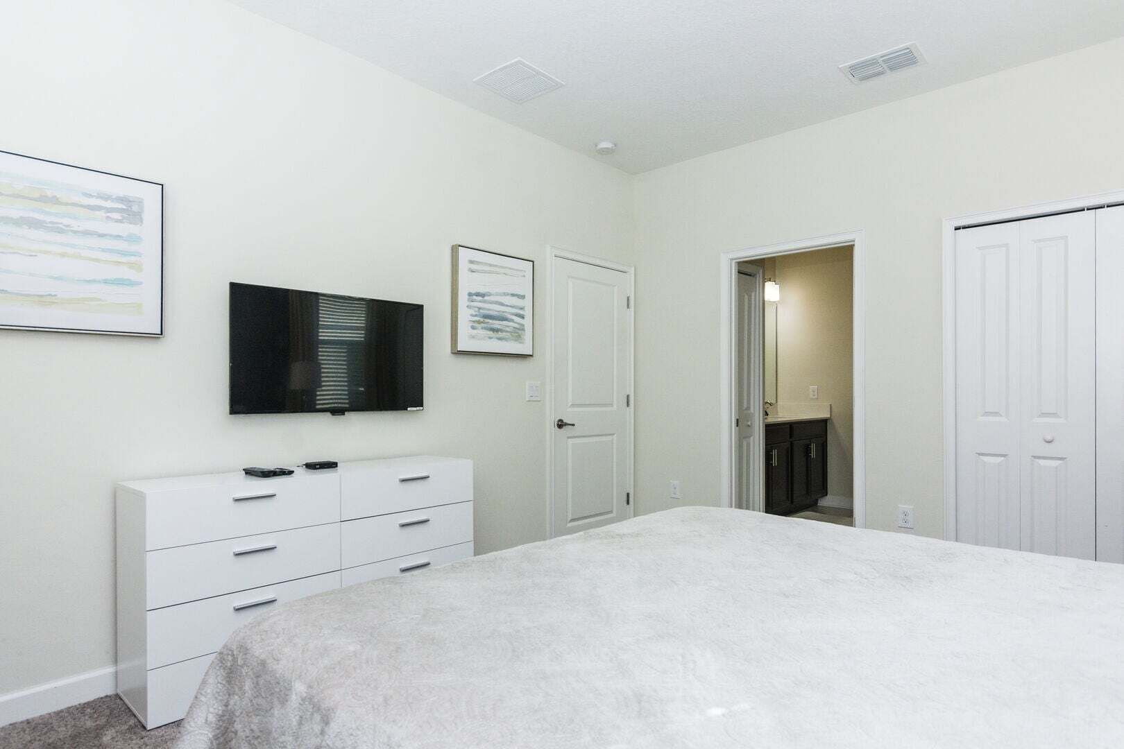 A bedroom with a bed, a dresser, a television, and a door leading to a bathroom.