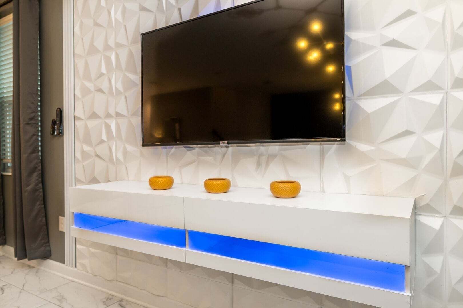 A modern living room with a flat-screen TV mounted on a textured wall, a white console table with blue lighting, and three decorative yellow vases.