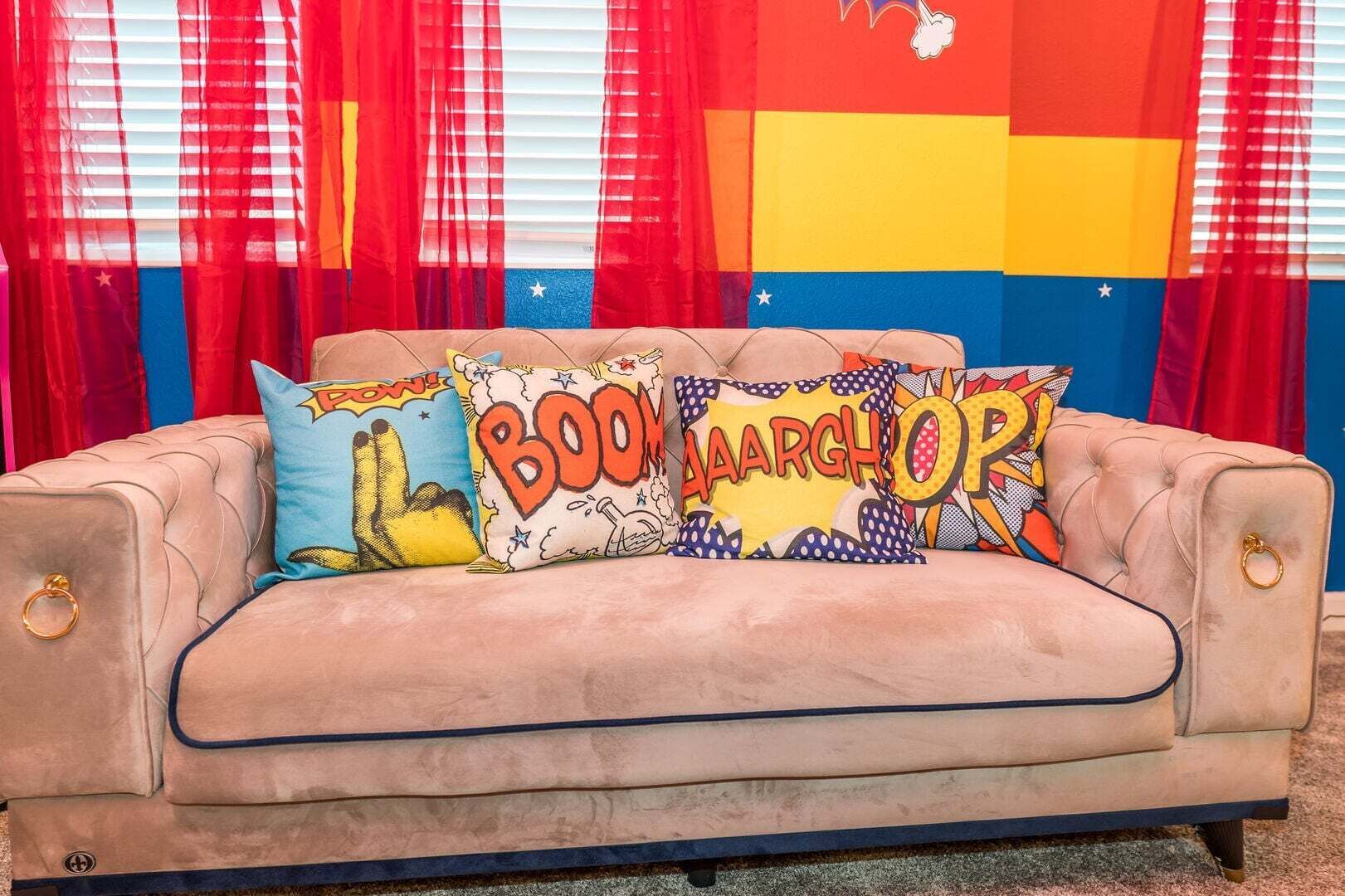 A beige sofa with three comic book-themed cushions, red curtains, and a colorful wall with stars.