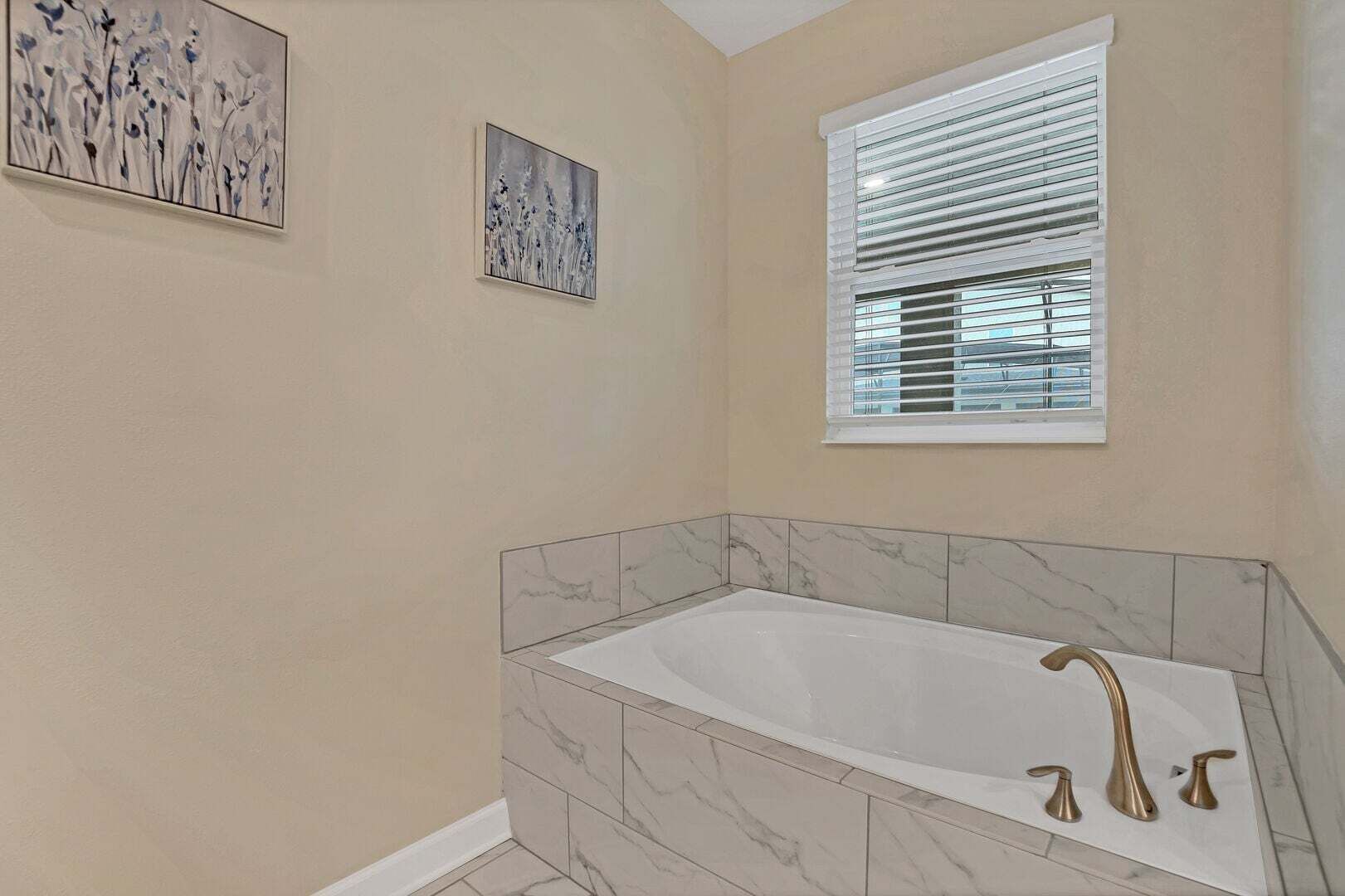 A bathroom with a marble tub, a window with blinds, and two wall-mounted artworks.