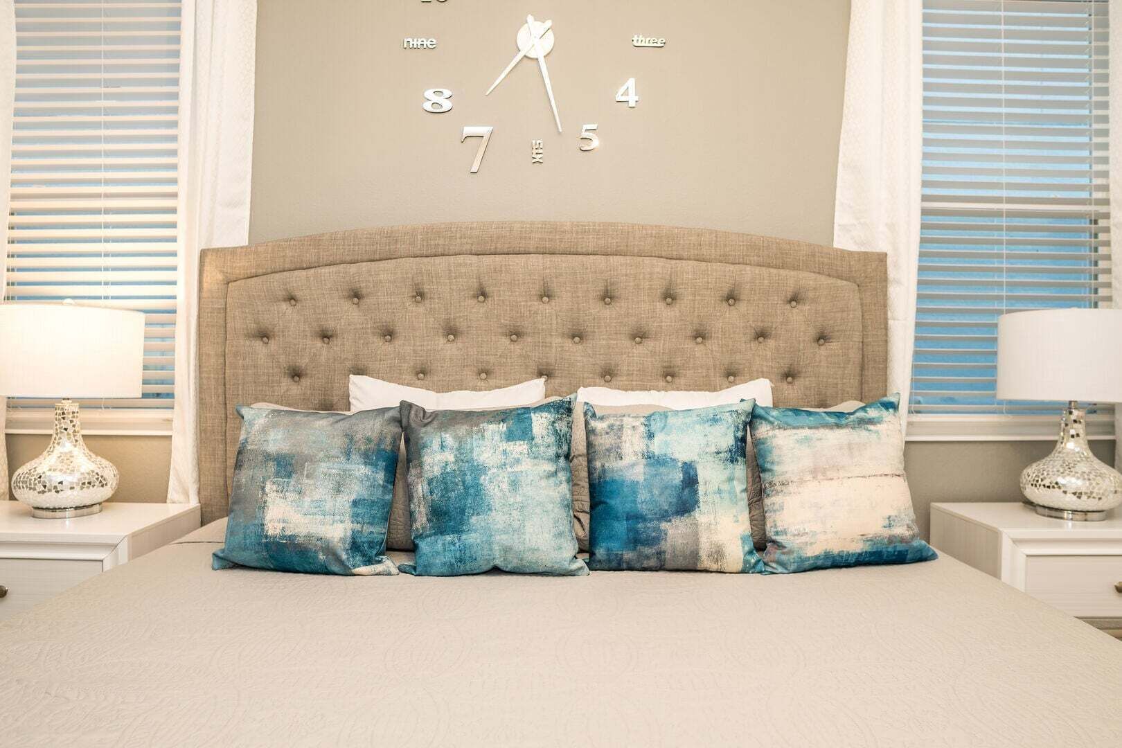 A bedroom with a tufted headboard, a bed with four pillows, a wall clock, and two bedside tables with lamps.