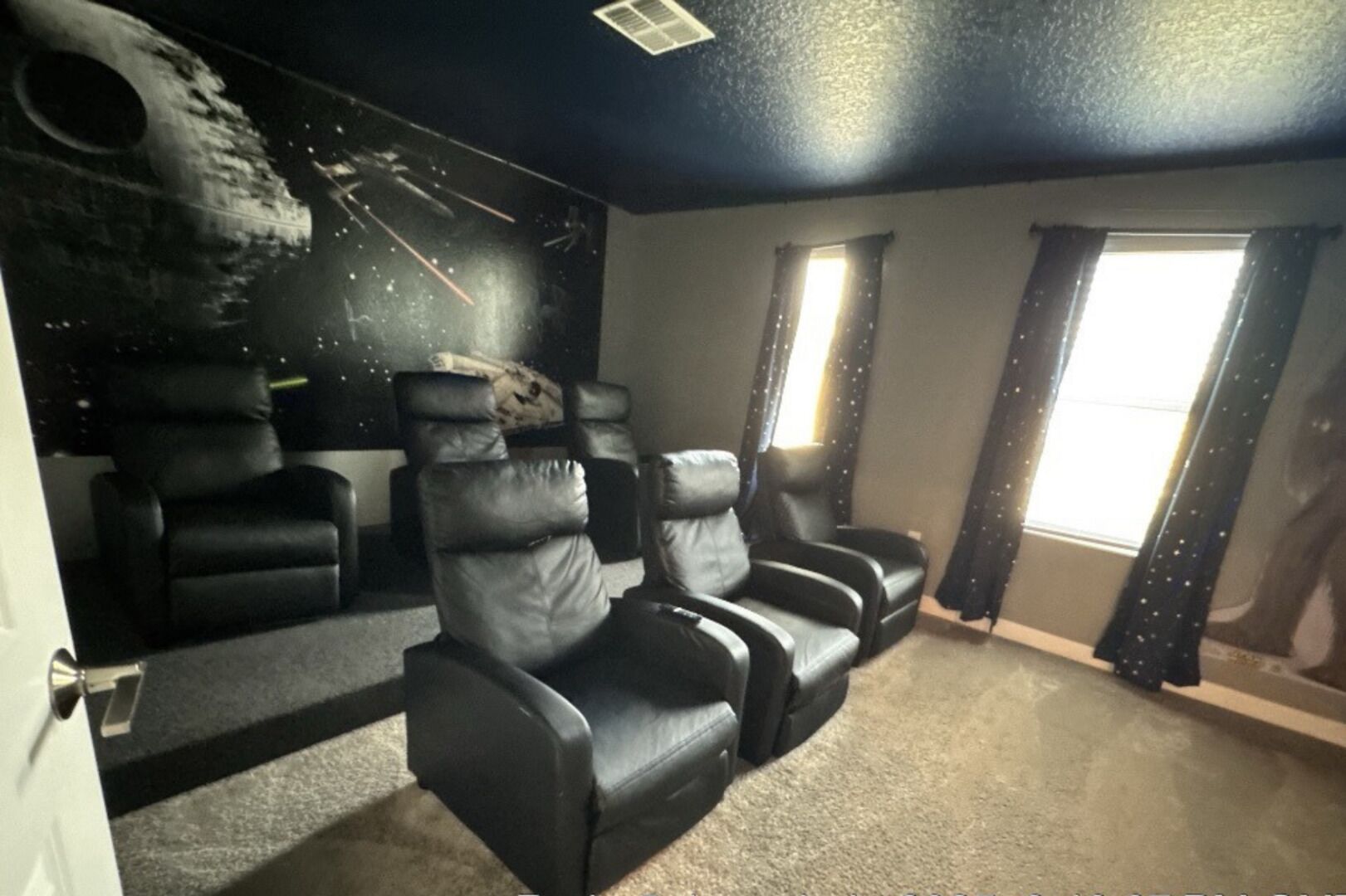 A room with a dark ceiling, a large window with curtains, and three black leather recliners.