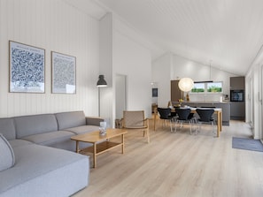 Ein modernes Wohnzimmer mit einem grauen Sofa, einem Holztisch und einem Essbereich mit Tisch und Stühlen. An der Wand hängen zwei gerahmte Kunstwerke, eine Pendelleuchte ist vorhanden, und im Hintergrund befindet sich eine Küche.