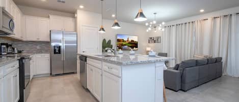 A modern kitchen with a central island, built-in appliances, pendant lighting, and a grey sofa in the adjacent living area.