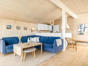 Ein Zimmer mit blauem Sofa, einem Holztisch, einem Korbstuhl und einer Küchenzeile mit weißen Schränken.