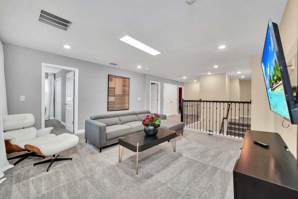 A spacious living area with a grey sofa, a coffee table with a vase of flowers, a flat-screen TV mounted on the wall, and a staircase leading to an upper level.