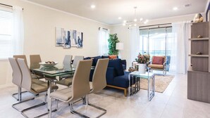 A modern living room with a glass dining table, comfortable seating, a sofa, and a large window. - Fantastic Home at Solterra Sol102 (Davenport)