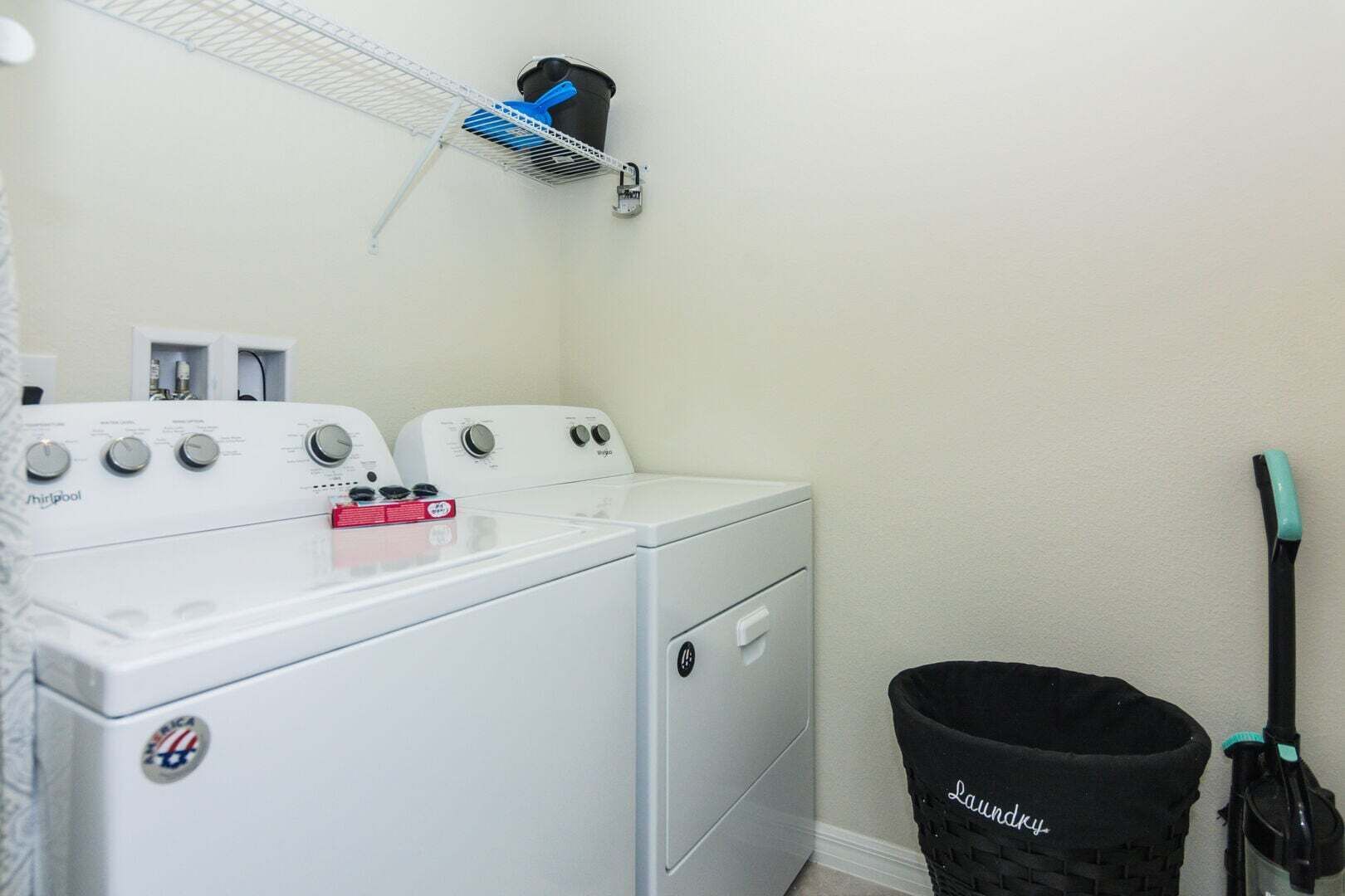 A laundry area with a front-loading washer and dryer, a shelf above the dryer, and a black laundry basket.