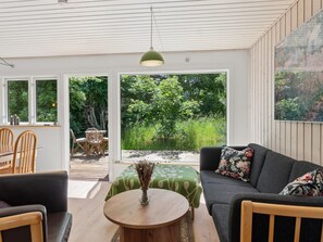 Ein modernes Wohnzimmer mit einer Couch, Sesseln, einem Tisch und Blick auf einen Garten durch große Fenster.
