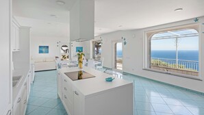 Un hotel moderno con una habitación amplia y ventana que ofrece vistas al mar, una isla central con un florero y un comedor con mobiliario blanco.