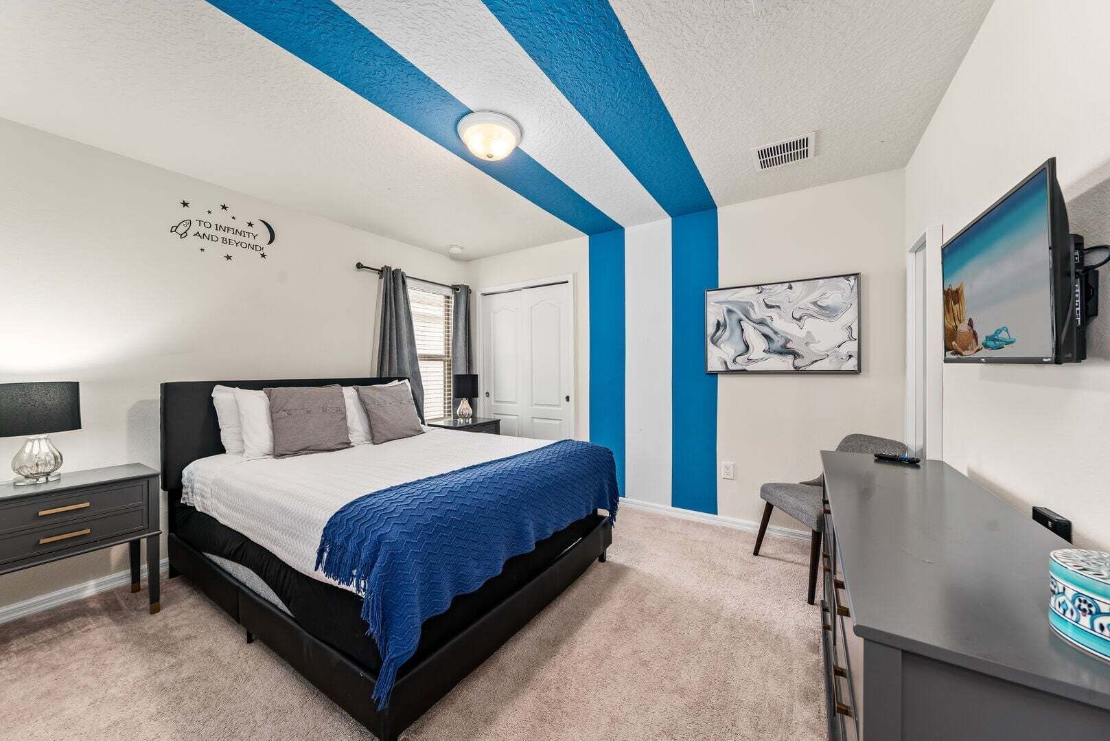 A bedroom with a bed, bedside table, desk, chair, artwork, and a door.