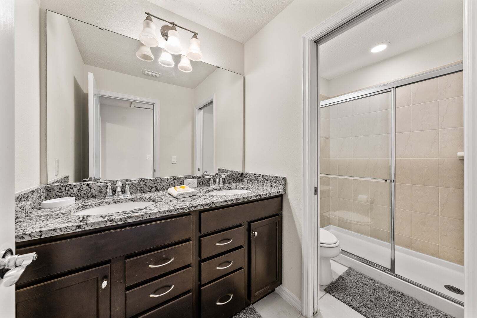 A bathroom with a granite countertop, dark cabinets, a large mirror, and a shower enclosure.