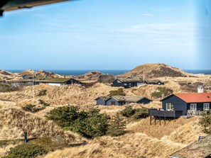 Eine Küstenlandschaft mit Häusern inmitten von Dünen, einem klaren Himmel und dem Meer im Hintergrund.