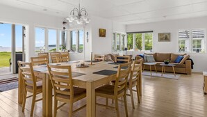 Quarto espaçoso com uma mesa de jantar de madeira e cadeiras, um sofá e grandes janelas com vista para o exterior.