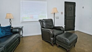 Een kamer met een zwarte leren bank, een leren fauteuil, een bijpassend poufje, een raam met jaloezieën, een deur en twee vloerlampen.