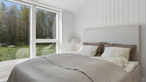Une chambre à coucher avec un grand lit, une table de chevet, une lampe et une vue sur une forêt, visible à travers une porte-fenêtre coulissante.