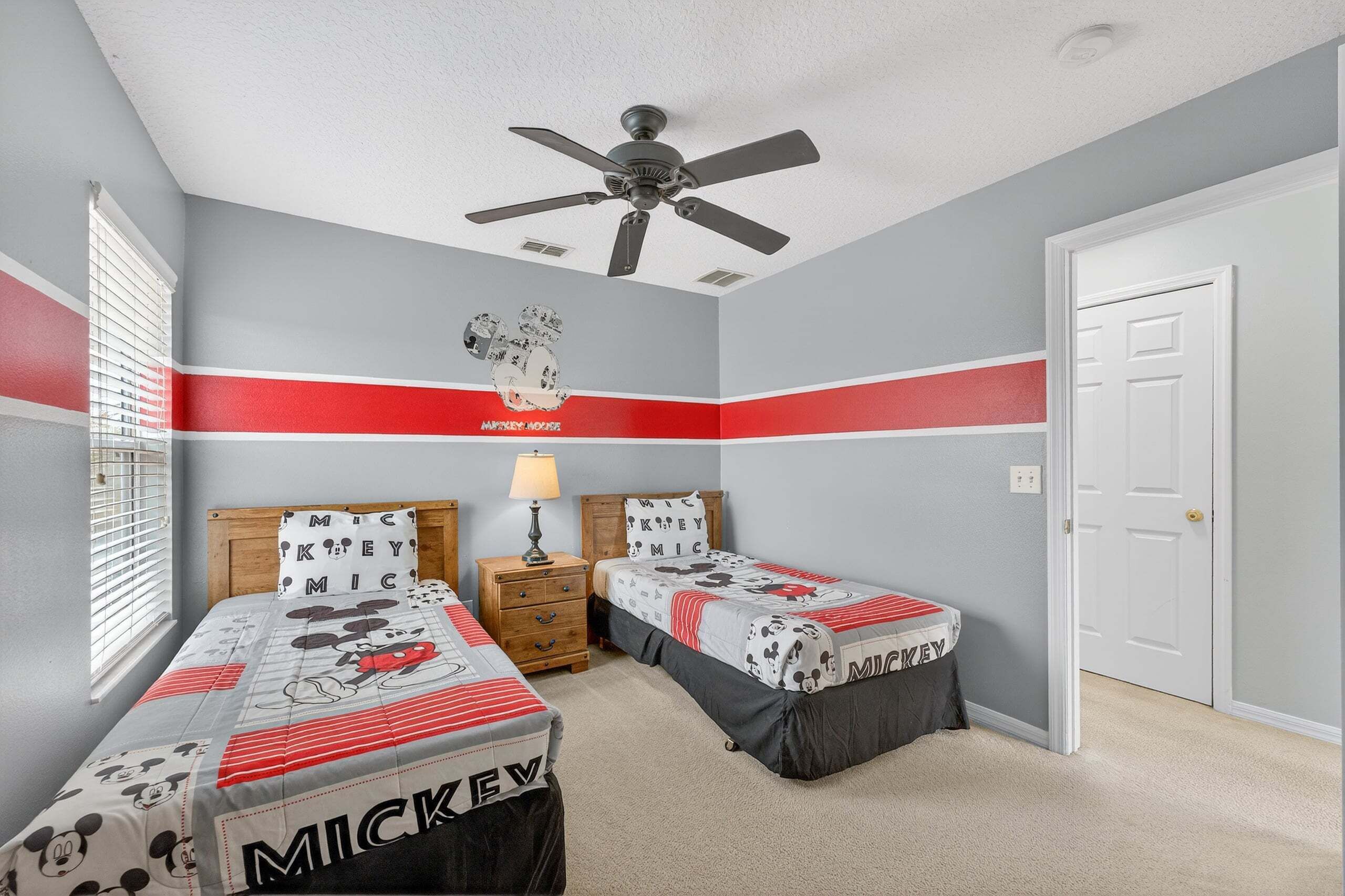 A bedroom with two beds, a ceiling fan, a window with blinds, and a door.