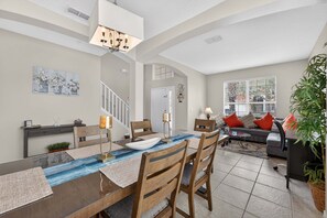 A dining area with a long table, chairs, and a chandelier. There is a staircase leading to another level, a sofa with cushions, and a large window with blinds. - Family Getaway: 6BR w/ Private Pool & Resort Access (Davenport)