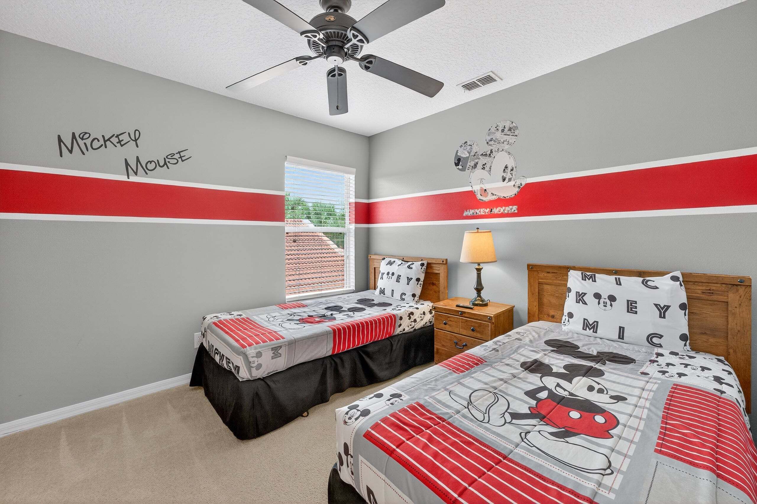 A bedroom with two beds, a ceiling fan, a window with blinds, and Mickey Mouse-themed bedding.