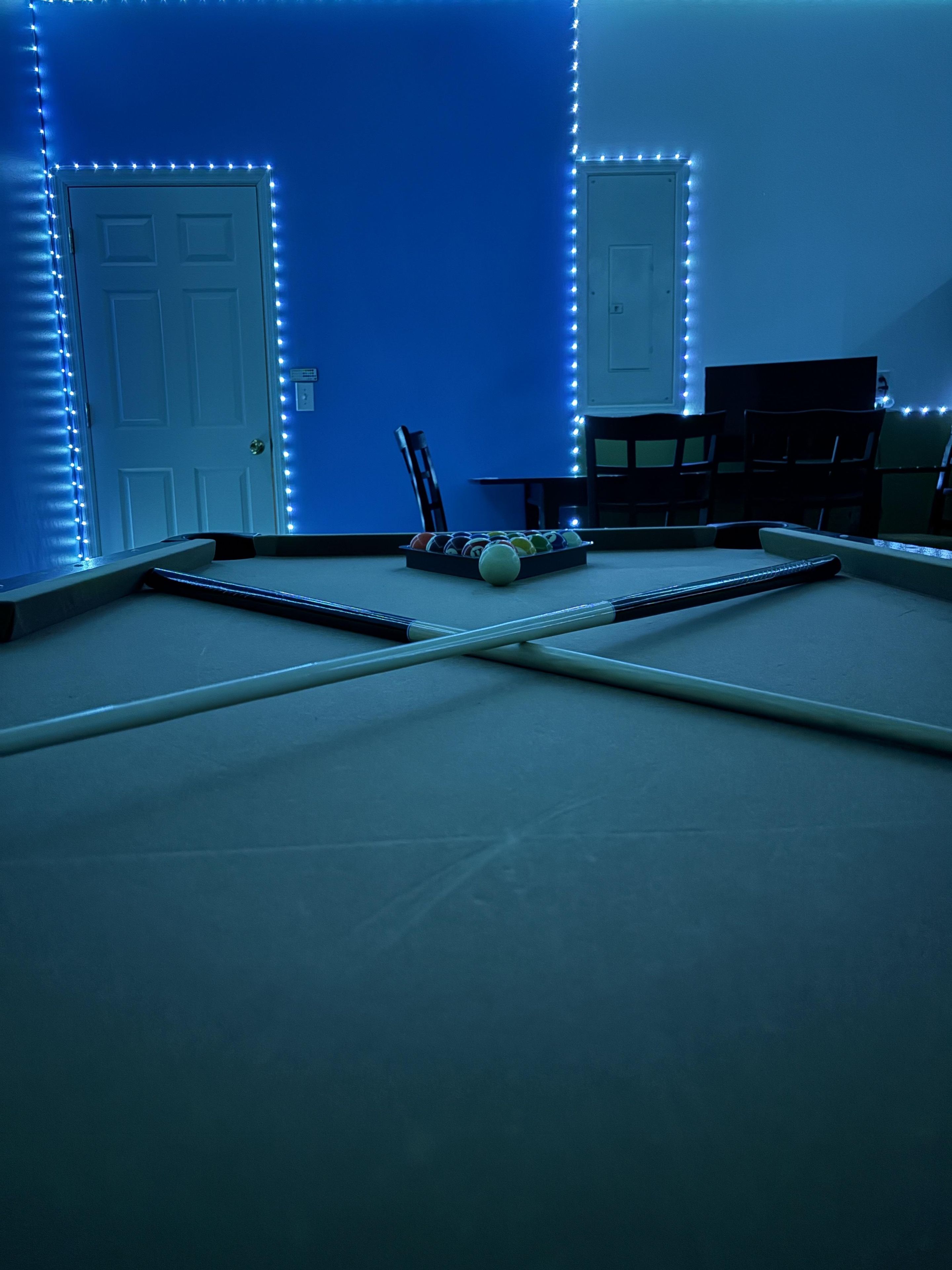 A pool table with a cue stick, a set of balls, and a chair in a room with blue walls and LED strip lighting.
