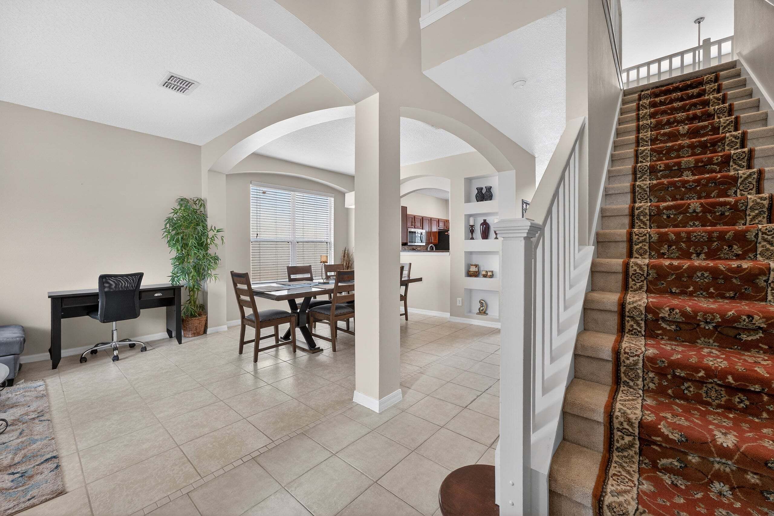 A spacious living area with a dining table, chairs, a desk, and a staircase with a patterned carpet.