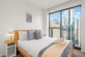 Ein modernes Hotelzimmer mit einem großen Bett, einem Nachttisch, einem Wandbild und Blick auf die Stadt durch ein großes Fenster.