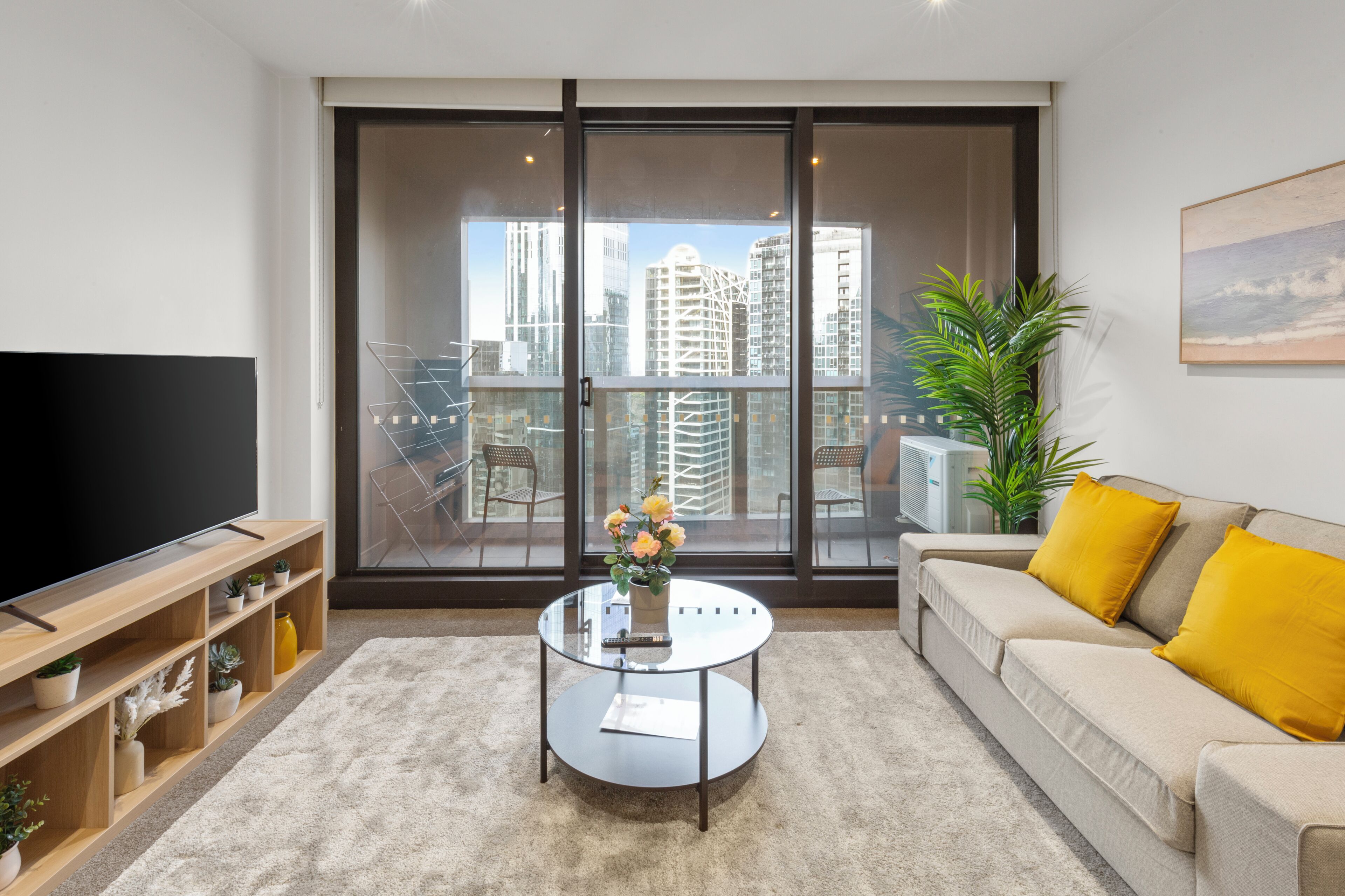 Una sala de estar moderna con un televisor de pantalla plana grande, una mesa de centro de vidrio con un florero, un sofá beige con cojines amarillos y vistas a la ciudad a través de ventanas de piso a techo.