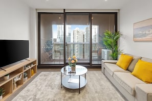 Ein modernes Wohnzimmer mit einem großen Flachbildfernseher, einem Glastisch mit Blumenvase, einem beigefarbenen Sofa mit gelben Kissen und einem Blick auf die Stadt durch raumhohe Fenster.