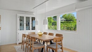 A dining room with a wooden table and chairs, white paneled walls, a large window with a view of greenery, and a cabinet with glass doors. - "Teofan" - 325m from the sea by Interhome (Ebberup)