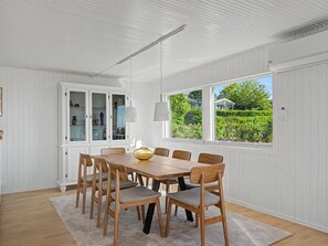 Ein Esszimmer mit Holztisch und -stühlen, weißen Wänden mit Paneelen, einem großen Fenster mit Blick auf Grünflächen und einem Schrank mit Glastüren.