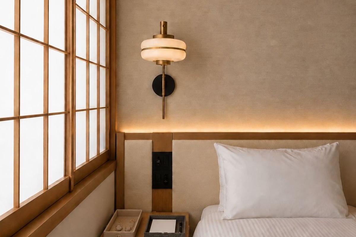 A hotel room with a bed, a nightstand, a wall-mounted lamp, and a window with a grid pattern.
