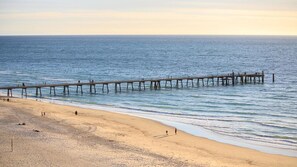 Ein Strand mit einem hölzernen Steg, der ins Meer führt, feinem Sand und blauem Himmel.