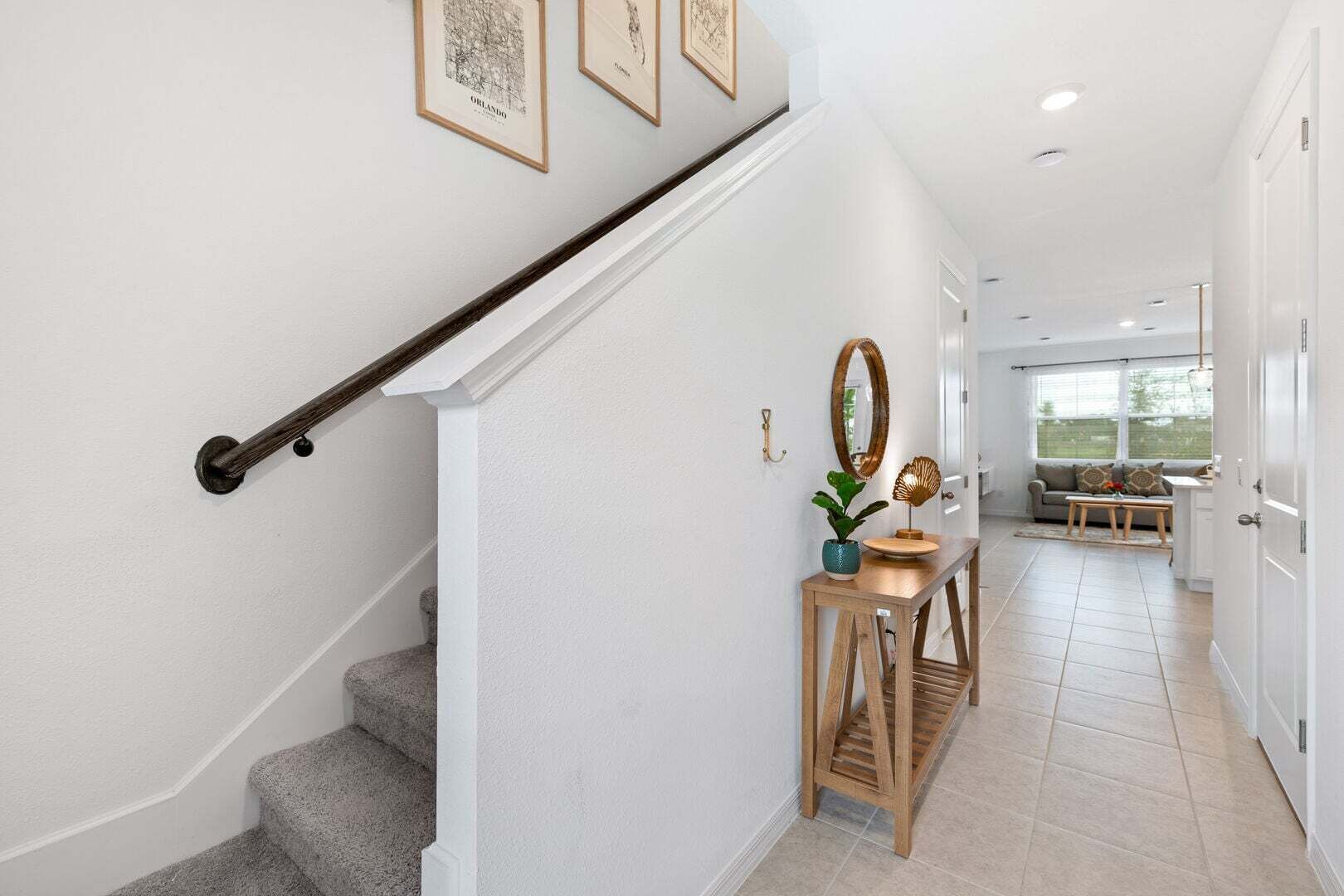 Una escalera con barandilla de madera, una pequeña consola de madera con un espejo y una planta, y vista hacia una sala de estar con un sofá y una ventana.