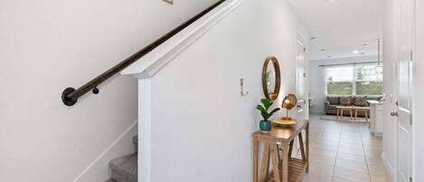Una escalera con barandilla de madera, una pequeña consola de madera con un espejo y una planta, y vista hacia una sala de estar con un sofá y una ventana.