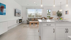A modern kitchen with white cabinetry, a dining area with a wooden table, and a living space with a sofa and TV. - Lake View Villa Sleeps 8! (Davenport)